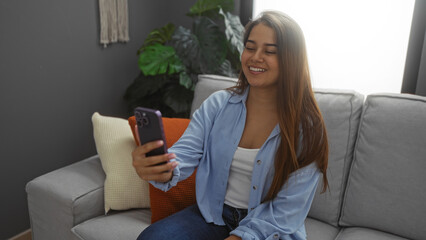 Obraz premium Woman talking on phone in a living room, wearing a blue shirt, with a smile, emphasizing her brunette hair and attractive features, creating a relaxed atmosphere in an indoor home setting