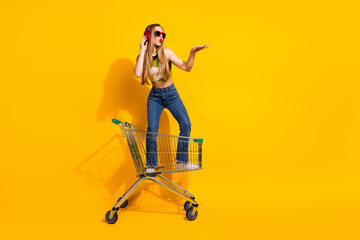 Fashionable young woman posing on a shopping cart against vibrant yellow background