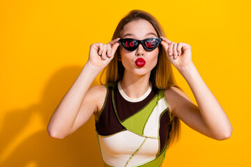 Young woman in trendy clothing posing playfully with sunglasses and a vibrant yellow background