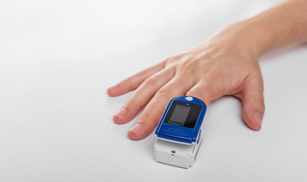 A close-up image of a person's hand with a blue and white pulse oximeter clipped onto their fingertip, used to measure oxygen saturation and heart rate.