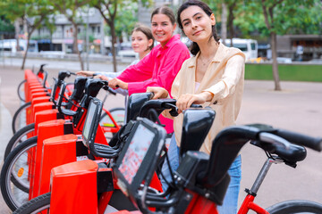 Obraz premium Happy young women renting bicycles in an urban park, smiling and preparing for a fun ride. Dressed casually, they enjoy a lively outdoor setting surrounded by trees and modern city buildings.