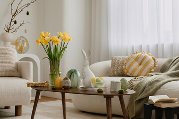 A bright and airy Easter-themed living space with yellow daffodils, a soft curved sofa, and woven rattan bunny decorations. The arrangement embraces modern rustic elegance.