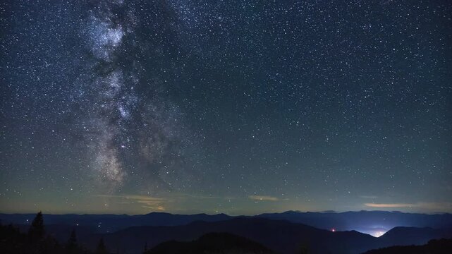 Time lapse video of vast night sky filled with stars and Milky Way spans above dark, undulating mountain range. Distant lights from scattered settlements add subtle glow, enhancing serene view.