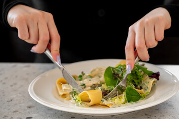 Close-up of a white plate with creamy pasta, fresh greens, and a fork and knife in hand, focusing...