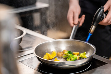 A professional chef uses a blowtorch to cook vegetables in a frying pan on a stovetop, with steam rising from broccoli, yellow bell pepper, and potatoes.