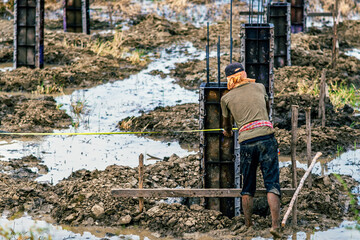 Male worker with Construction of concrete pillars in paddy field. Formwork with reinforcement bar for pad footing. Construction of pad footing foundation. Metal formwork for concrete foundations.