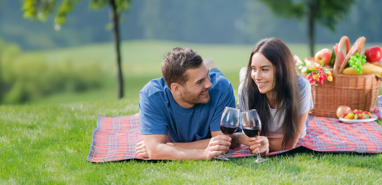 Young happy smile couple enjoys a picnic grassy field, lying on plaid blanket. Man and woman look at each other, hold glasses of red wine drink, against summer park nature background. Banner image