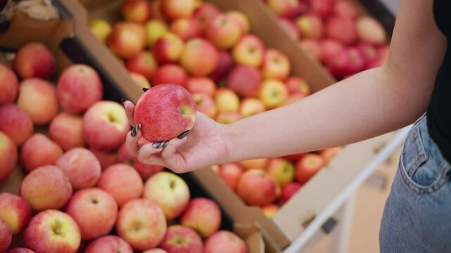 Close-up of woman picking a red apple from a box of apples, observing it briefly before placing it back into the box in a grocery store setting