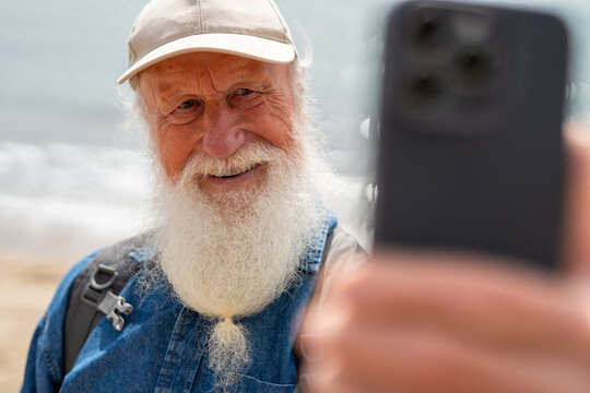happy elderly man with a long white beard, wearing a beige cap and denim shirt, takes a close-up selfie with his smartphone while standing on a sunny beach with ocean waves in the background