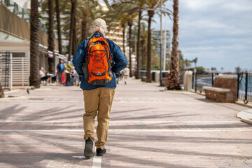 An elderly man with a white beard and a cap, wearing a denim shirt and carrying a bright orange backpack, strolls along a palm tree-lined promenade on a sunny day