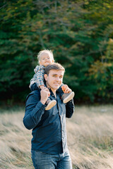 Fototapeta premium Smiling dad with a little girl on his shoulders stands in the park