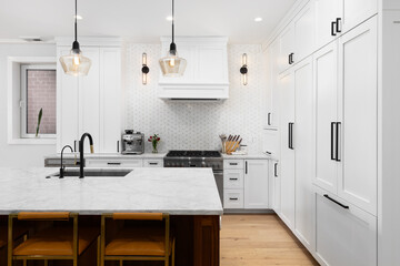 A white kitchen detail with white cabinets and marble countertop, bar stools sitting at a large...