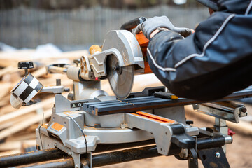 Close up of a worker using a miter saw to cut wood, with gloves and jacket for safety. Background shows blurred wooden planks and a fence.