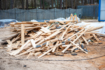 A construction site features wooden planks with metal brackets, sandy ground, a blue temporary structure, and a tall wooden fence near trees.