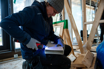 A worker uses a power drill on a white duct piece for a ventilation system in a partially constructed house with wooden framing and large windows.