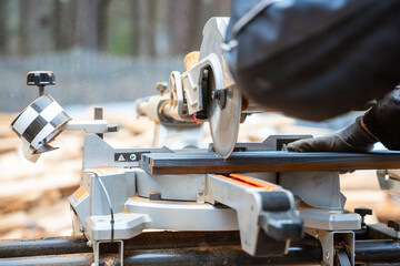 Close up of a circular saw cutting material on a miter saw setup. A gloved operator adjusts the equipment, with blurred trees in the background.