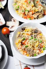 Couscous is a dish made from wheat grains (semolina) or millet. Lean couscous with mushrooms, corn, peas, herbs, tomatoes. Side view. Flatlay. Couscous on a light background. Couscous in a white plate