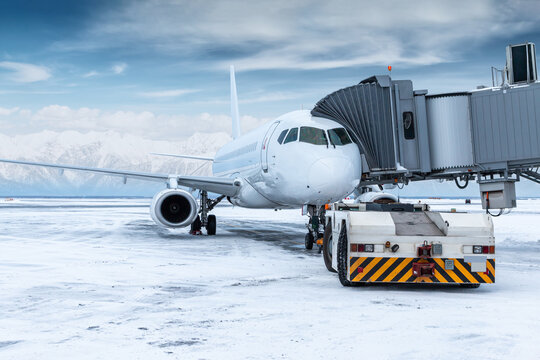 The tow tractor approaches the white passenger airplane at the aerobridge on the background of high snow covered mountains