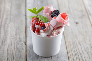 Delicious rolled ice cream in a cup, garnished with fresh berries, a chocolate wafer stick, and fruit toppings, served on a rustic wooden table