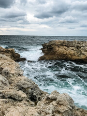 A picturesque rocky shoreline where waves crash powerfully against the large rocks on an overcast, cloudy day filled with a gray sky and a feeling of natures raw beauty
