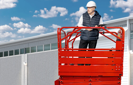 construction supervisor in a hard hat and vest stands on a red scissor lift, inspecting an industrial building exterior with a clipboard under a bright sky - Powered by Adobe