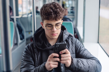 Thoughtful teenage boy using smartphone on public transport, young man with glasses in winter...
