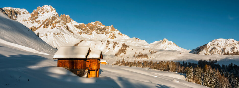 A wooden cabin sits in a snowy landscape with mountains in the background under a clear blue sky.  Pian di Fuciade, Pale di San Martino, Dolomites, Trentino, Italy