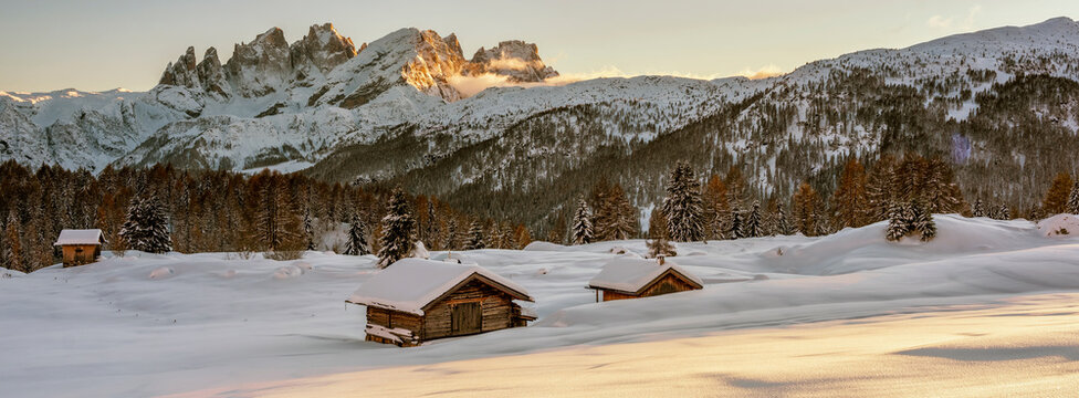 Snowy mountain landscape with two small wooden cabins in the foreground. The mountains are partially covered in trees and snow, with the peaks catching the golden light of the setting sun. 