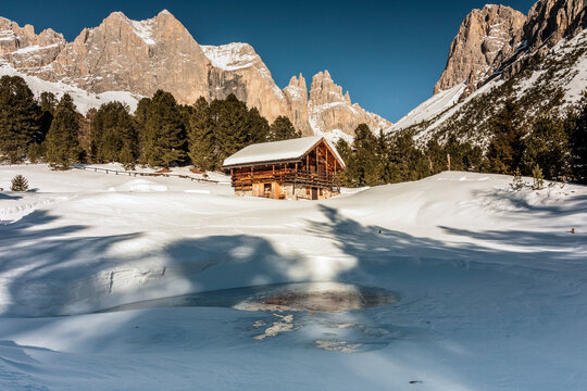 A wooden cabin sits in a snowy landscape, framed by towering mountains and evergreen trees under a clear blue sky. A partially frozen pond reflects the scene.