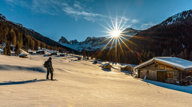 A hiker walks across a snowy field toward wooden cabins in a mountain valley. The sun shines brightly behind the mountains. Valle di San Nicol&ograve;., Val di Fassa, Dolomites, Trentino, Italy