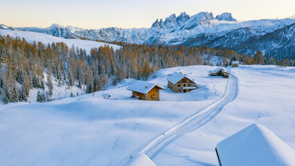 Snowy mountain landscape with wooden cabins in Pian di Fuciade, Trentino, Dolomites. A path leads through the snow to the cabins, with a forest of golden trees and jagged mountains in the background. 