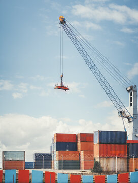 A large industrial crane lifts a red cargo platform with a worker standing on it at a shipping port. Stacked colorful freight containers and a barbed-wire fence are visible