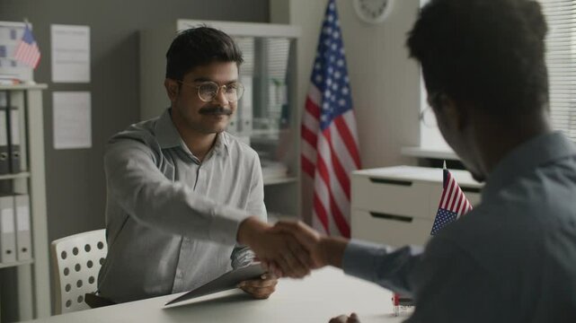 Immigration officer returning visa application form to Indian man, then shaking his hand after approval during formal interview at US embassy