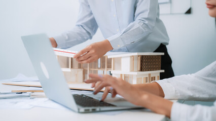 Closeup image of professional male architect hand using ruler to measure house model length while young coworker using laptop to analyzed data on meeting table with house model. Immaculate.