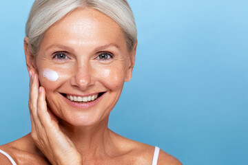 Beauty portrait. Anti age skin care cream tretments. Smiling mature woman with applying moisturiser under her face, showing a relaxed skincare routine against a blue background.