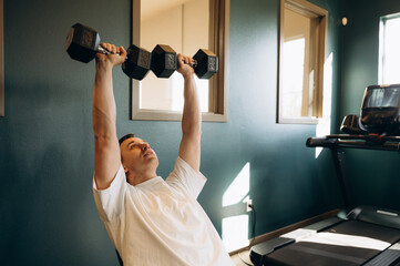 In a spacious gym, a man takes a position on a bench press. He grips the dumbbells tightly in his hands, tensing the muscles in his shoulders and arms.