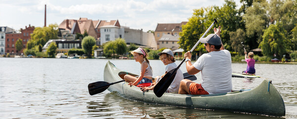 Man with two children enjoy having fun paddling canoe rent across lake river water wearing bright orange life vests hot sunny summer day. Healthy recreational activity lifestyle vacation trip journey