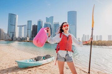 Young woman enjoys kayaking in front of Abu Dhabi skyline, holding a paddle and a waterproof bag