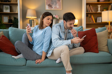 Obraz premium Woman spying man's phone while sitting back-to-back on couch at home, both using mobile phones