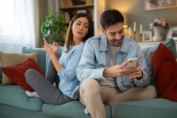 Woman spying man's phone while sitting back-to-back on couch at home, both using mobile phones