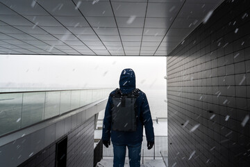 A man with a backpack in winter clothes stands in a modern arch of a building in snowfall. Light in the background, a dark building and a man in a hood with his back to the camera.