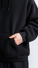 Person wearing black fleece hoodie puts their hand in the front pocket against a clean background, showcasing the soft texture and garment construc...