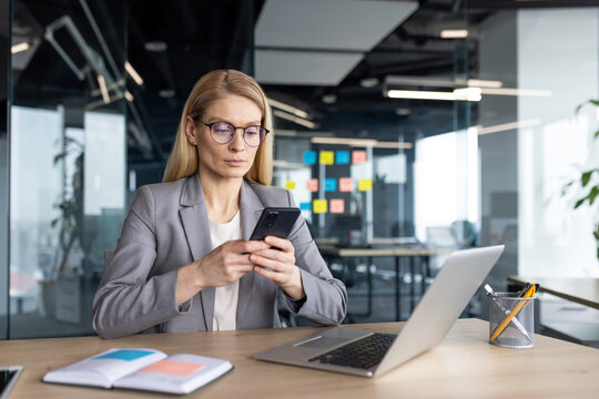 A businesswoman in a jacket is using her mobile phone while sitting at a desk in a modern office setting.