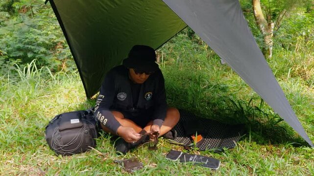 A male adventurer is cooking sauce to make a burger under a flysheet in the dense forest. The concept of bushcraft camping in a dense pine forest for adventure and cooking with simple equipment