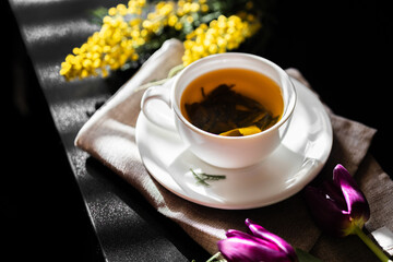White cup with green tea on a dark background in sunlight. White porcelain cup with saucer. Yellow flowers nearby. Large green tea leaves unfolded in the cup.