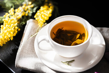 White cup with green tea on a dark background in sunlight. White porcelain cup with saucer. Yellow flowers nearby. Large green tea leaves unfolded in the cup.