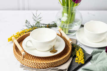 Still life of white porcelain cups and teapot, tulips in a glass vase, mimosa flowers, light background. Tea party. Spring tea in good weather.