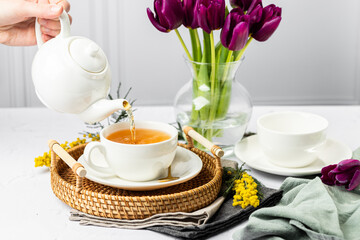 Green tea is poured from a white teapot into white porcelain cups. In the background are tulips in a glass vase, mimosa flowers, light background. Tea drinking.
