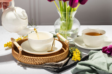 Green tea is poured from a white teapot into white porcelain cups. In the background are tulips in a glass vase, mimosa flowers, light background. Tea drinking.