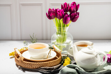 Green tea in a white porcelain cups and teapot, tulips in a glass vase, mimosa flowers, light background. Tea party.
